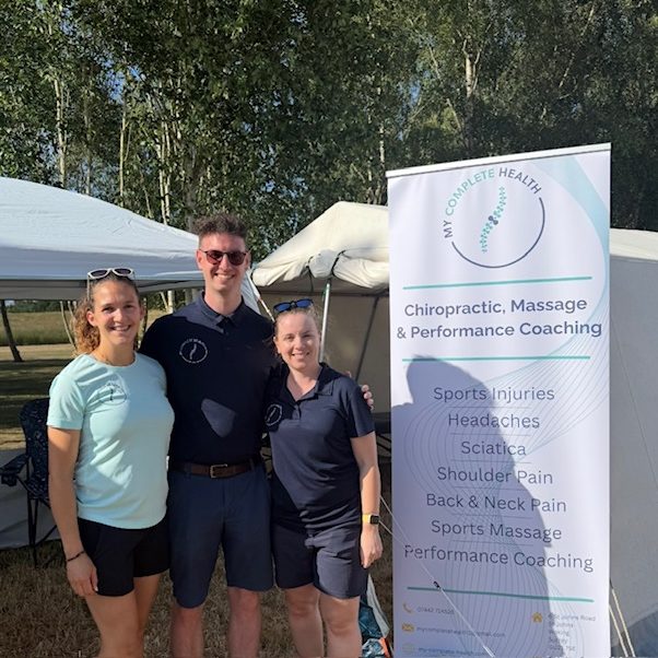 The team at My Complete Health. The male chiropractor is in the middle with two female members of the team either side of him. They all have their arms around each other. Next to them is a poster advertising the services the clinic offers.